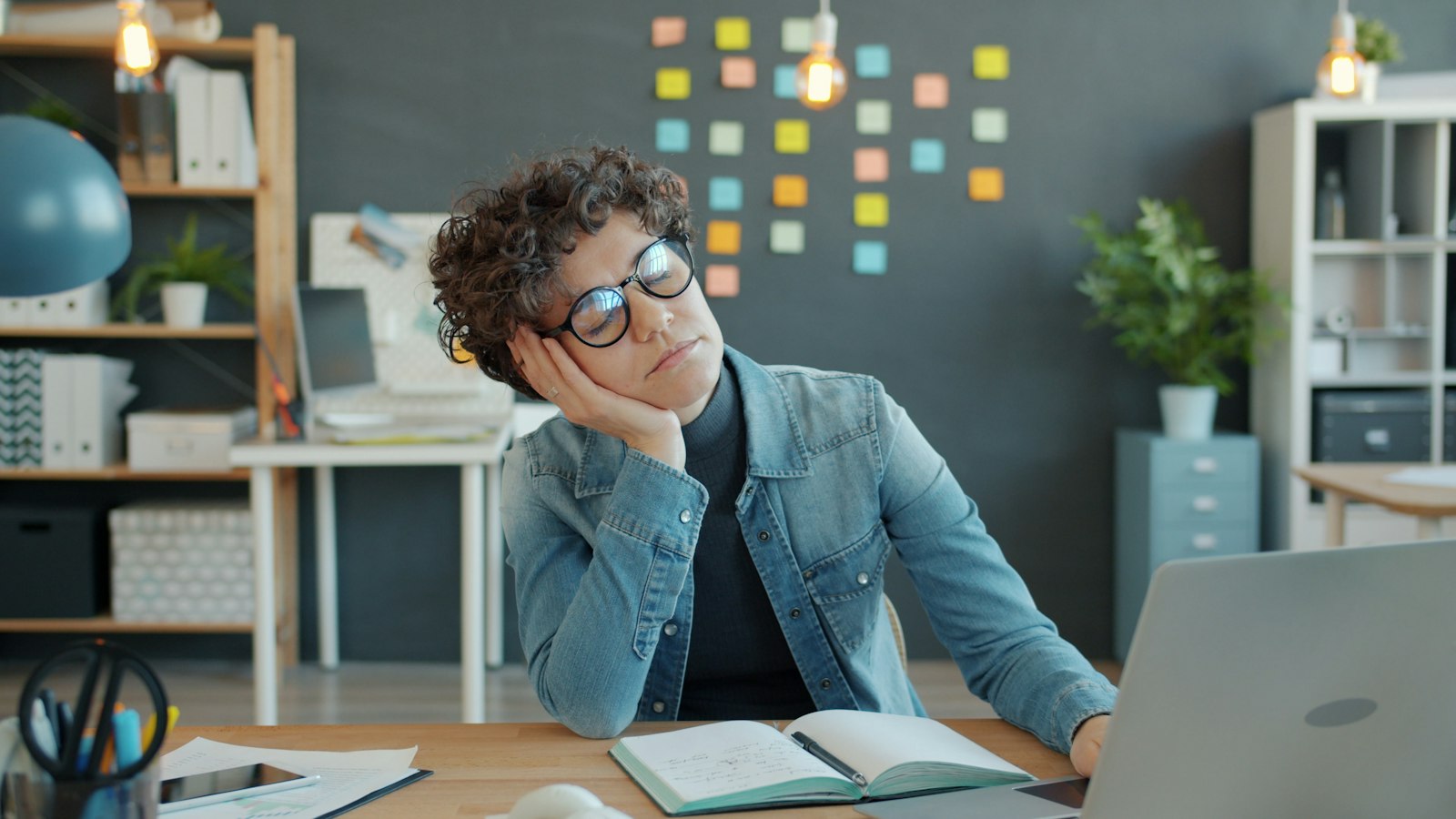 Adult at a desk with a laptop and notebook, resting their head on one hand and looking distracted